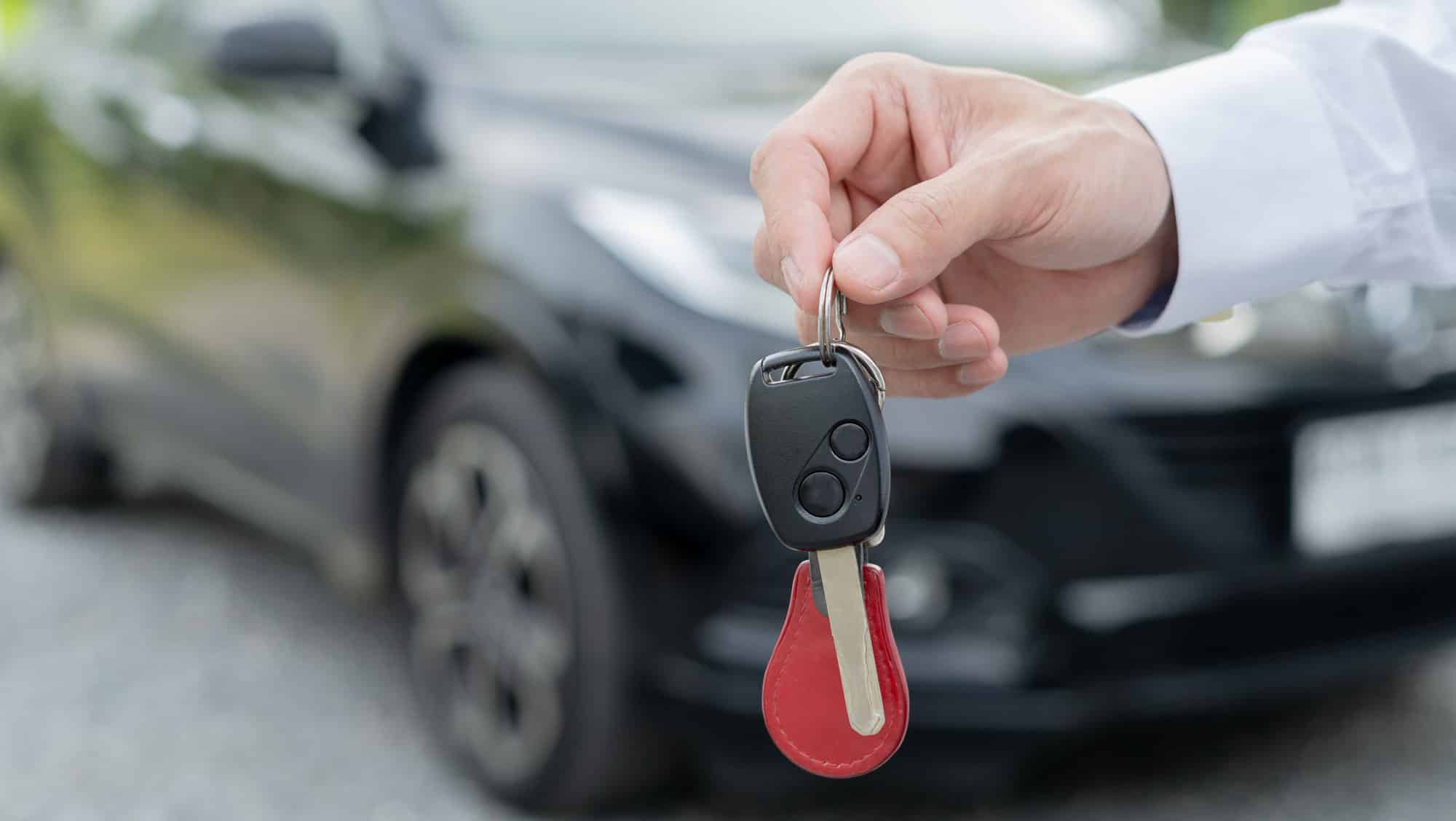 A man's hand holding car keys in the foreground with a blurred out car in the background