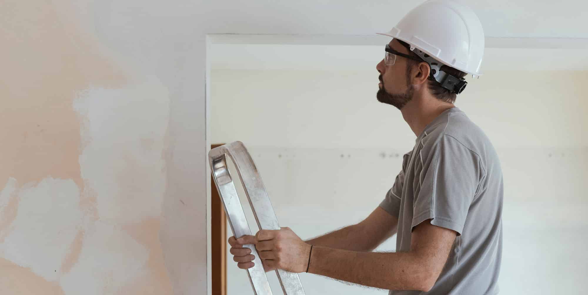 A man with a white hard hat on a ladder, looking at drywall