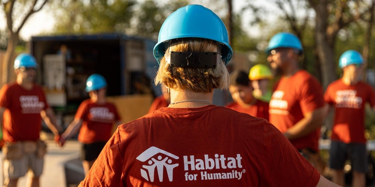A group of Habitat for Humanity volunteers with blue hard hats on, focused on the back of one volunteer, showing the logo on the back of her shirt